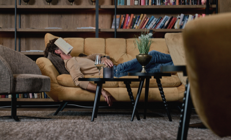 A man is lying on a yellow sofa in the bar area of the hotel with a book on his head and a glass of wine next to him