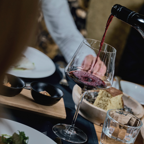 A glass of red wine standing at the dinner table, into which a glass from the bottle is being poured